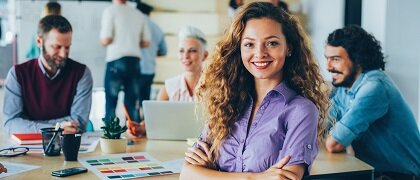 Junge Menschen sitzen um einen Tisch in Büro und eine Frau mit verschränkten Armen steht im Vordergrund