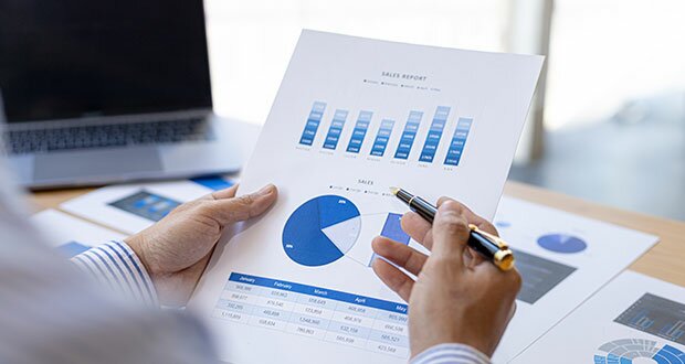 Man sieht ein Papier mit verschiedenen blauen Diagrammen. Dahinter ein Schreibtisch mit einem Laptop und weiteren Papieren.